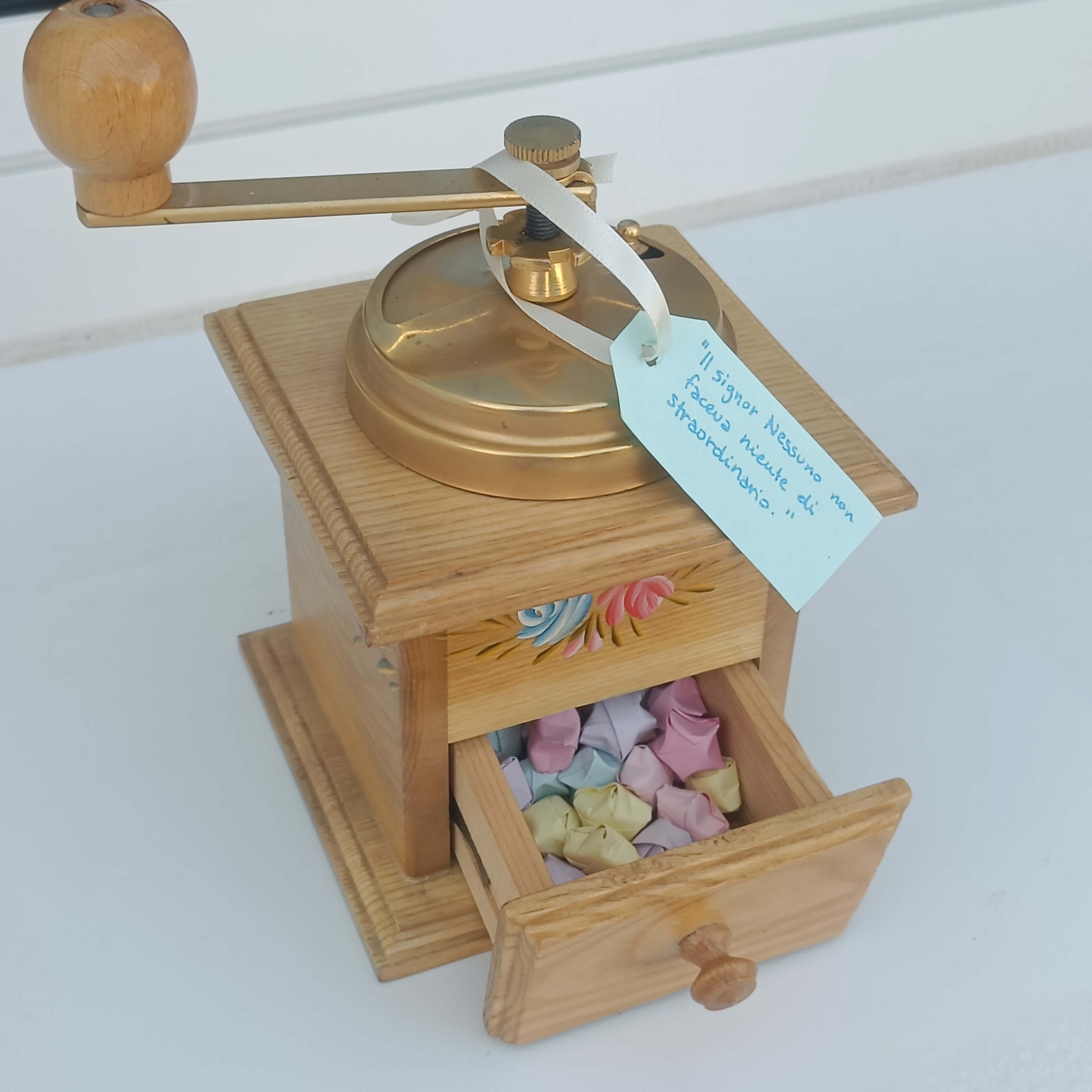 A picture of an old wooden grinder. Its drawer is filled with colored paper stars. There's a handwritten tag on it reading: 'Il signor Nessuno non faceva niente di straordinario.'