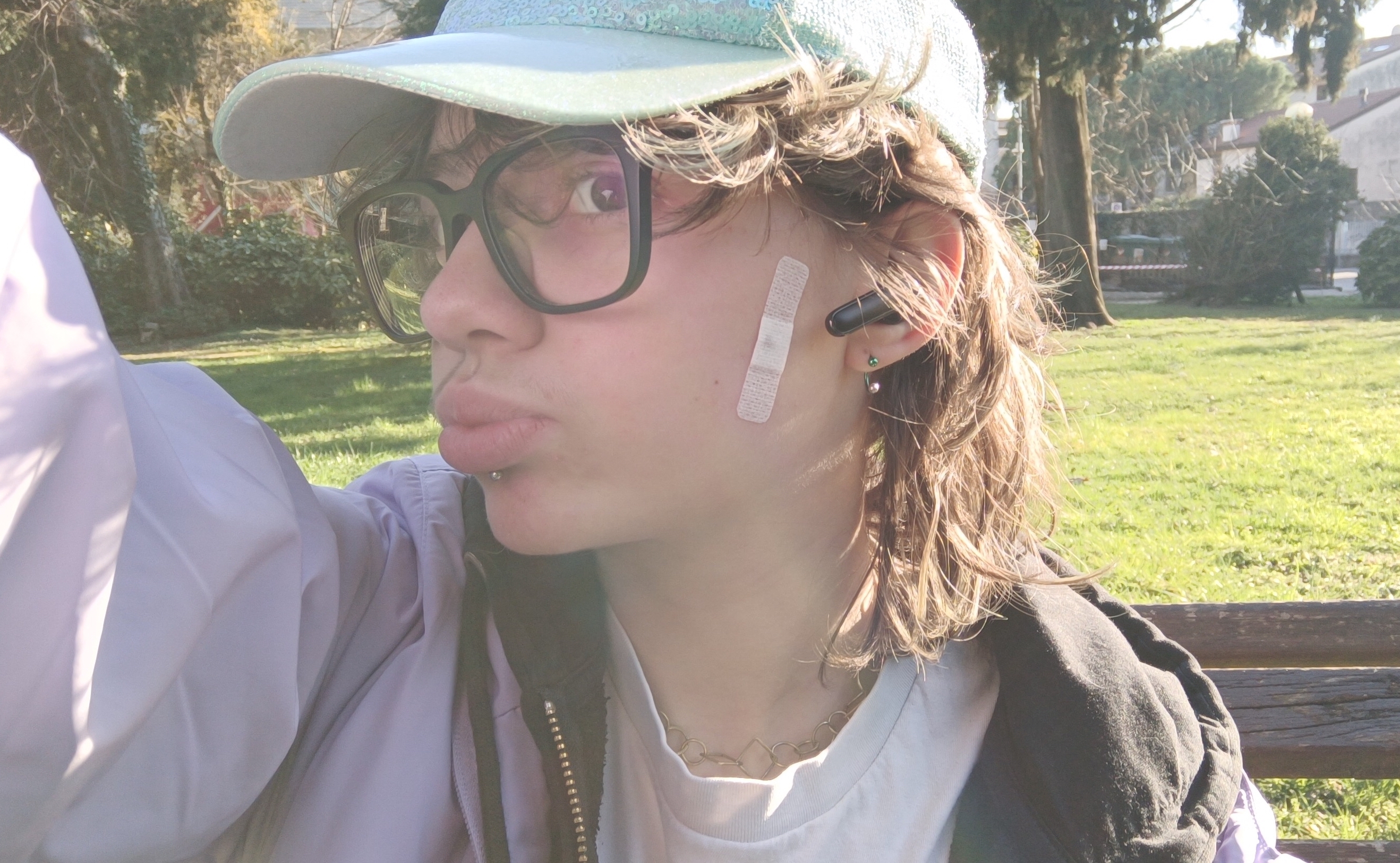 A selfie of Elliot in a park. He is a white man and he has brown medium length hair. He is seen from the bust up and he is doing a kissy face. He is wearing a white shirt,a black hoodie and a lilac jacket. He has a shiny light blue cap and a labret piercing. He has a white bandaid on his cheek.