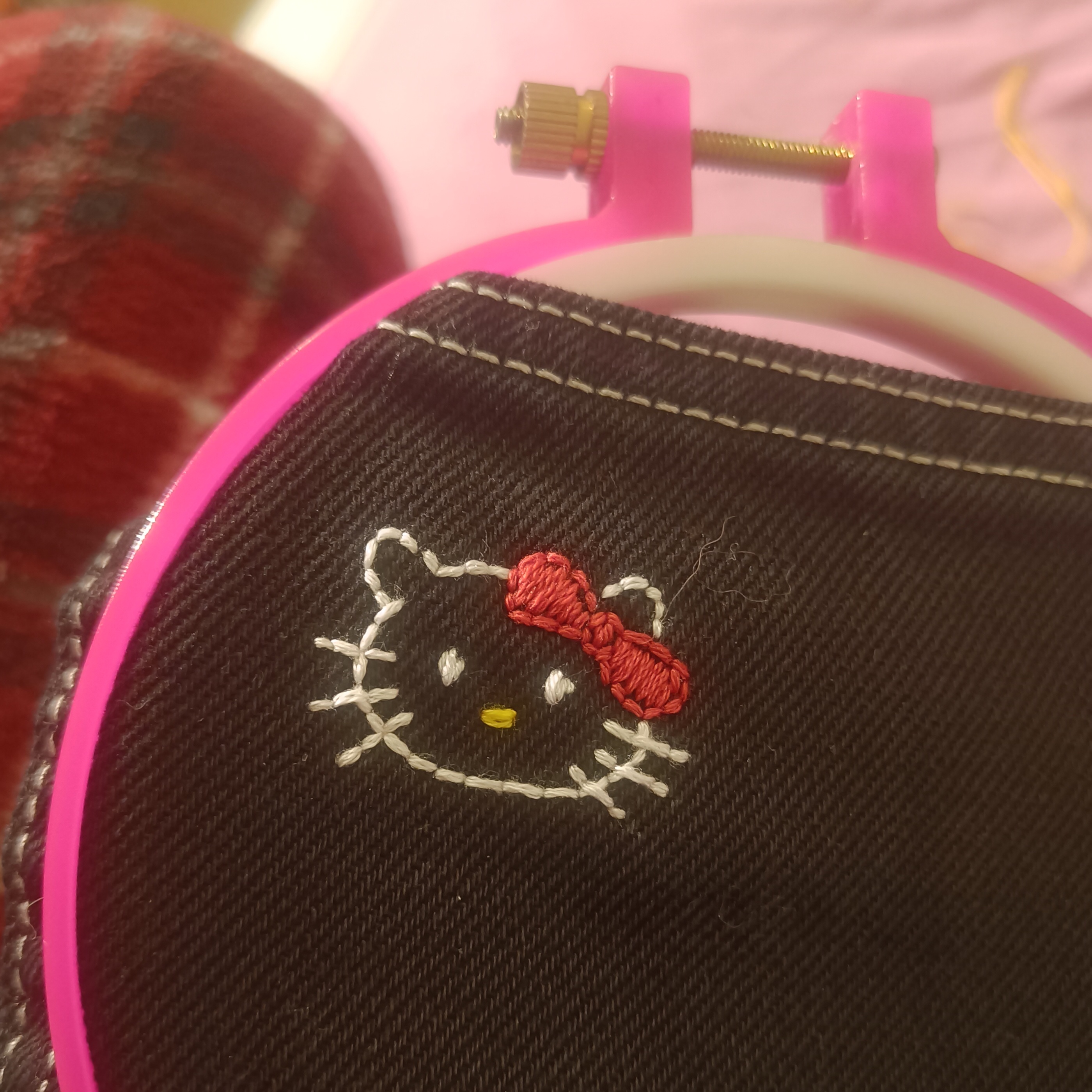A picture of an embroidery hoop placed on the back pocket of a pair of black jeans, with the face of Hello Kitty hand-embroidered on it.